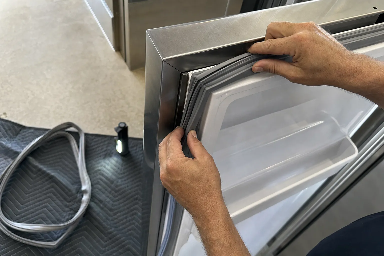 Hands seating a new gasket into the channel of a stainless appliance door, old gasket coiled on the mat